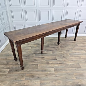 May include: A long, rectangular wooden table with a dark brown finish and a smooth, flat surface. The table is supported by six straight, tapered legs that extend to the floor, providing a stable base for the elongated tabletop structure.