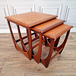 May include: A set of three nested tables crafted from medium-toned wood. Each table features a rectangular top with rounded edges and curved, continuous side supports that form a distinct arch shape, allowing the smaller tables to slide underneath.