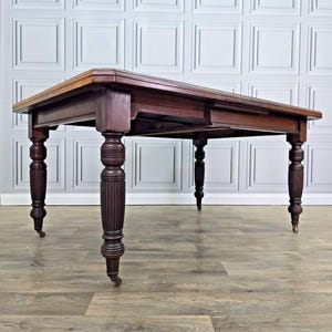 May include: A rectangular dark brown wooden dining table with a smooth top surface and four thick, vertically fluted legs. Each leg features a rounded, decorative carved section at the top and is supported by a small metal caster wheel at the base.