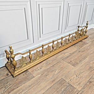 May include: A long, horizontal brass fireplace fender featuring a decorative railing with vertical spindles and alternating fan-shaped motifs. The metal structure has a polished gold-toned finish and includes two taller finials at each end.