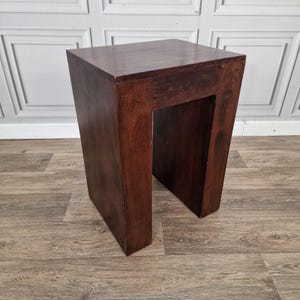 May include: A dark brown wooden side table with a rectangular top and a U-shaped base. The piece features a smooth, solid construction with visible wood grain patterns throughout the surface, standing as a singular, sturdy furniture item.