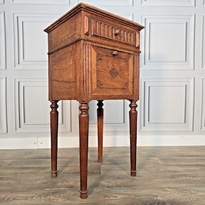 May include: A tall, rectangular wooden side table featuring a single top drawer with vertical fluting and a lower cabinet door. The cabinet door is decorated with carved floral motifs. The piece stands on four slender, turned wooden legs.