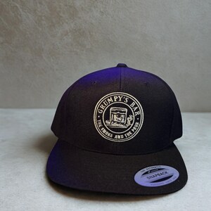 Puede incluir: Gorra snapback negra con un logotipo circular blanco que dice "Grumpy's Bar" y "The Smoke and The Pour". La gorra tiene una visera plana y un cierre a presi&oacute;n. Hecha de un material resistente.