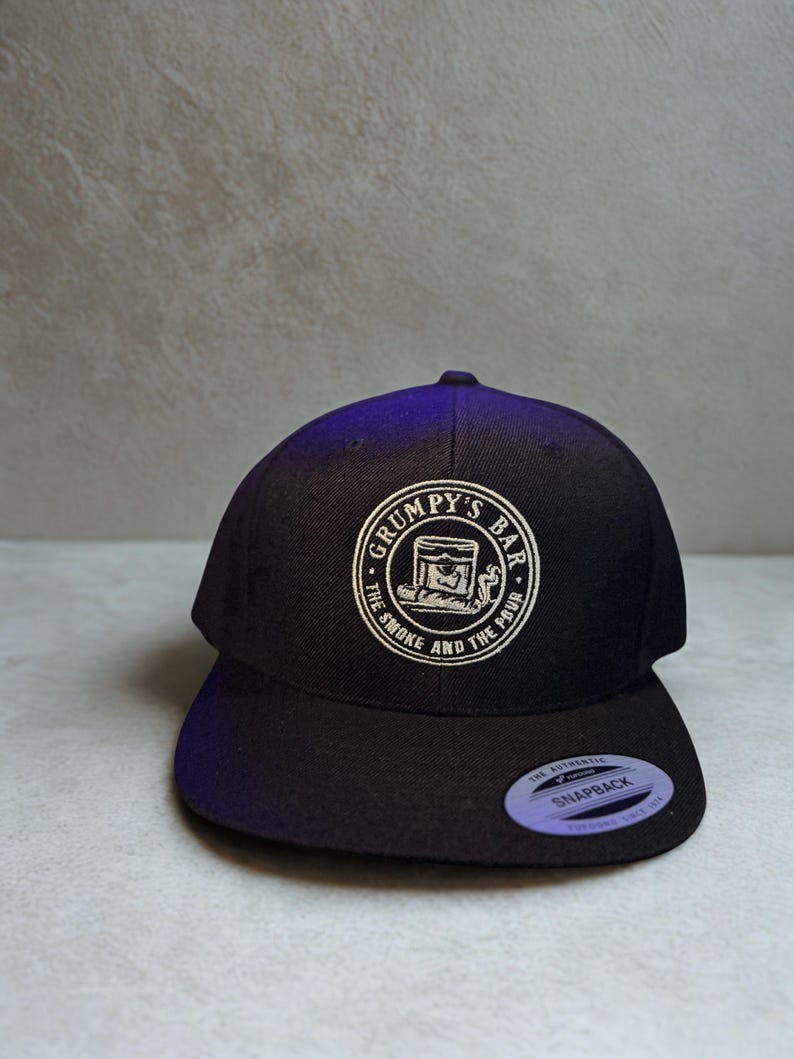 Puede incluir: Gorra snapback negra con un logotipo circular blanco que dice "Grumpy's Bar" y "The Smoke and The Pour". La gorra tiene una visera plana y un cierre a presi&oacute;n. Hecha de un material resistente.
