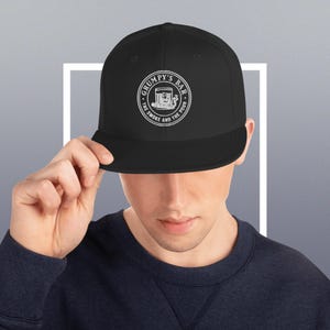 Könnte beinhalten: Schwarze Baseballkappe mit flachem Schirm. Die Kappe zeigt ein weißes kreisförmiges Logo mit dem Text "Grumpy's Bar" und dem Satz "The Smoke and The Pour". Das Logo enthält eine Cocktail-Illustration.