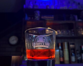 Puede incluir: Un vaso bajo lleno de un líquido rojo-anaranjado, probablemente un cóctel, descansa sobre una superficie oscura. El vaso tiene la marca "Grumpy's Bar" y "The Smoke & The Pour". Un letrero de neón es visible en el fondo.