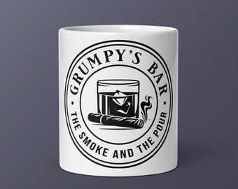 Puede incluir: Taza de cerámica blanca con un diseño gráfico negro. El diseño presenta un vaso de whisky con hielo, un cigarro y el texto "Grumpy's Bar - The Smoke and The Pour" en un arreglo circular. La taza es cilíndrica.