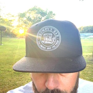 Puede incluir: Gorra negra de ala plana con el texto "GRUMPY'S BAR" en un dise&ntilde;o circular. El dise&ntilde;o incluye una ilustraci&oacute;n de un vaso y las palabras "THE SMOKE AND THE POUR". La gorra se usa al aire libre frente a un campo de hierba.