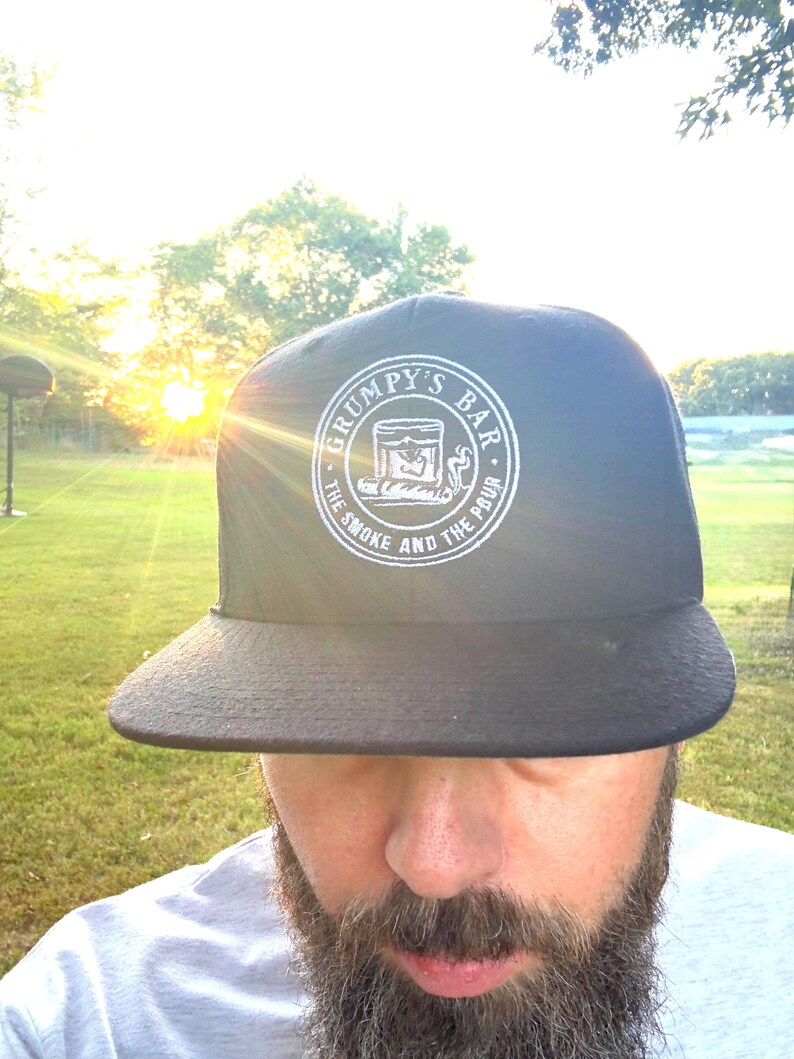 Puede incluir: Gorra negra de ala plana con el texto "GRUMPY'S BAR" en un dise&ntilde;o circular. El dise&ntilde;o incluye una ilustraci&oacute;n de un vaso y las palabras "THE SMOKE AND THE POUR". La gorra se usa al aire libre frente a un campo de hierba.