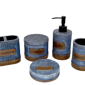 Puede incluir: Un juego de baño de cinco piezas con diseño de mezclilla. El juego incluye un porta cepillos de dientes, un tarro de algodón, un dispensador de jabón, un jabonera y un vaso. Cada pieza tiene detalles y etiquetas de cuero marrón.