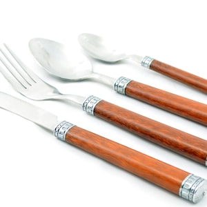 Puede incluir: Un juego de cuatro utensilios de acero inoxidable: un cuchillo, un tenedor y dos cucharas. Cada pieza tiene un mango de madera marrón claro con detalles metálicos plateados. Dispuestos sobre un fondo blanco.