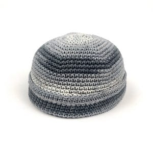 May include: A crocheted skull cap in shades of grey. The hat features a rounded crown and a folded brim. The colour transitions from light grey at the top to darker greys at the brim. The hat is made of a soft, textured yarn.