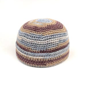 May include: A round, crocheted beanie hat featuring horizontal stripes in muted shades of light blue, beige, tan, and dark brown. The hat is constructed with a dense, textured stitch pattern and includes a folded brim detail around the base.