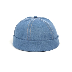 Könnte beinhalten: Eine blaue Denim-Docker-Mütze mit einer abgerundeten Krone aus sechs Paneelen, die durch sichtbare hellbraune Nähte verbunden sind. Die Mütze hat einen dicken, umgeschlagenen Rand, der die Basis umschließt und eine schmale, eng anliegende Form bildet.
