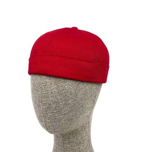 Puede incluir: Un gorro estilo docker de color rojo brillante con una corona redondeada y un borde grueso doblado. El gorro está confeccionado con una tela de color sólido con costuras visibles a lo largo de los paneles, creando una silueta estructurada.