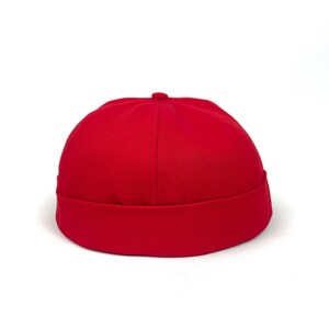 Puede incluir: Una gorra roja lisa sin visera que presenta una corona redondeada y un borde grueso doblado en la base. El sombrero está confeccionado con un tejido duradero con una textura visible y un pequeño detalle de botón en el centro superior.