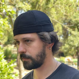 Op de afbeelding: Een zwarte skullcap-hoed. De hoed is gemaakt van een donkere, effen stof en heeft een ronde kroon en een kleine rand. De hoed wordt op het hoofd gedragen. De achtergrond is wazig, met groen gebladerte.