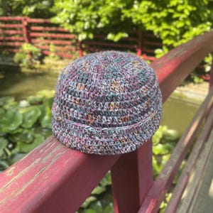 May include: A crochet beanie hat featuring a multi-coloured yarn blend with flecks of blue, purple, pink, and orange. The hat has a rounded crown and a thick, folded brim, showcasing a textured stitch pattern throughout the entire construction.