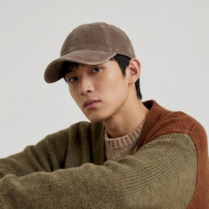 May include: A brown corduroy baseball cap with a curved brim. The cap is a solid color and has a classic six-panel design. The cap is worn by a person wearing a green and brown sweater.