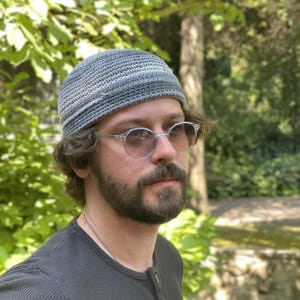 May include: A grey crochet beanie with a textured, ribbed brim. The hat features a tight, uniform stitch pattern in varying shades of grey, creating a subtle gradient effect. The beanie has a rounded crown and a folded edge that sits close to the head.