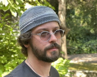 Puede incluir: Un gorro tejido gris con un diseño a rayas. La persona lleva gafas redondas con montura transparente y una camisa gris oscuro. El fondo presenta follaje verde y un tronco de árbol.