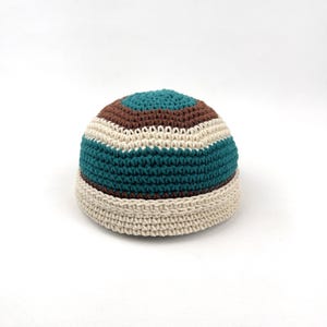May include: A crocheted beanie hat with a striped pattern. The hat features bands of teal, brown, and off-white, with a beige brim. The hat is a dome shape and is made of a soft yarn.