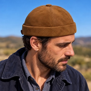 Puede incluir: Un gorro docker de ante marrón con una corona redondeada, un diseño sin ala cosido y un pequeño botón en el centro. El gorro tiene un aspecto estructurado con detalles de costuras visibles a los lados y un borde doblado hacia arriba.