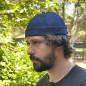 Könnte beinhalten: Eine einfarbige marineblaue Dockermütze mit abgerundeter Krone und einem dicken, umgeschlagenen Rand. Die Mütze besteht aus einem glatten, matten Stoff mit sichtbaren Nähten, die von der Mitte ausgehen und eine strukturierte, schirmlose Form bilden.