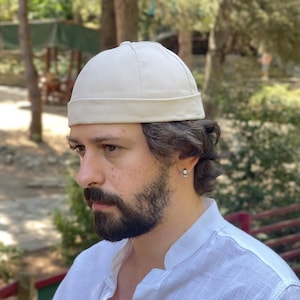 Puede incluir: Un gorro tipo docker de color blanco roto con una corona redondeada y un borde grueso doblado. El gorro está confeccionado con un tejido liso y resistente con detalles de costuras visibles a lo largo de los paneles, creando una silueta limpia.