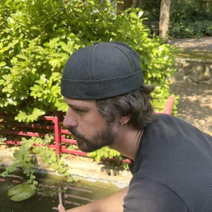 May include: A dark charcoal grey docker cap featuring a rounded crown, a thick folded brim, and visible orange stitching along the seams. The hat is constructed from a textured fabric and has a structured, brimless design that sits high on the head.