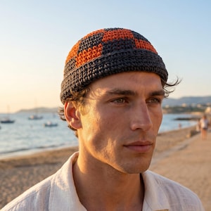 Puede incluir: Un gorro de ganchillo con un patrón geométrico en naranja y gris oscuro. El sombrero lo lleva una persona en una playa. El sombrero es un accesorio elegante para el uso informal.