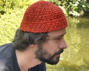 Puede incluir: Un sombrero de ganchillo de color óxido con una copa redondeada y un ala ligeramente más ancha. El sombrero está hecho de un hilo texturizado y parece estar hecho a mano. El sombrero lo lleva una persona con barba.