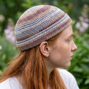 Op de afbeelding: Een gestreepte, gehaakte beanie in tinten bruin, beige, blauw en paars. De hoed is handgemaakt en heeft een nauwsluitend ontwerp. De beanie is geschikt voor koel weer en voegt een vleugje handgemaakte charme toe aan elke outfit.