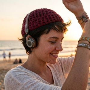 Puede incluir: Una persona con un gorro de ganchillo burdeos y auriculares en una playa al atardecer. Los auriculares tienen detalles de cuero marrón y cascos plateados. La persona lleva una camisa de manga larga de color claro y una pulsera que dice "SUNFEST '24".