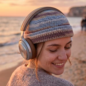 May include: A person wearing a multi-coloured crocheted beanie and silver and brown over-ear headphones. The headphones are around the person's neck. The background is a beach with the sea and a sunset.