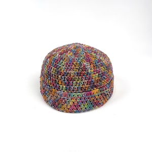 May include: A multi-coloured crocheted beanie hat. The hat is made with a variety of yarns in shades of pink, blue, yellow, and green, giving it a textured, handmade look. The hat has a round shape with a folded brim.