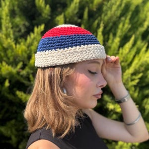 Könnte beinhalten: Eine gehäkelte Mütze mit einem Color-Blocking-Design, bestehend aus einem roten Oberteil, einem marineblauen Mittelstreifen und einem dicken, cremefarbenen Rand. Die Mütze ist in einem sichtbaren Strukturmuster gefertigt und eng anliegend.
