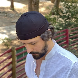 Puede incluir: Un gorro tipo docker negro liso con una copa redondeada, un pequeño botón forrado de tela en la parte superior central y un borde grueso doblado. El gorro está confeccionado en tela lisa y mate con costuras visibles desde el centro.