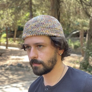 Puede incluir: Un gorro de ganchillo multicolor con una corona redondeada. El sombrero presenta una mezcla de hilos amarillos, verdes, azules y marrones. El sombrero lo lleva una persona con barba y pelo oscuro.