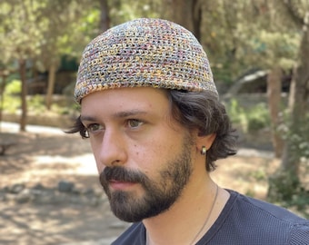 Puede incluir: Un gorro de ganchillo multicolor con una corona redondeada. El sombrero presenta una mezcla de hilos amarillos, verdes, azules y marrones. El sombrero lo lleva una persona con barba y pelo oscuro.