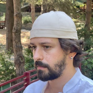 May include: A solid off-white docker cap featuring a rounded crown and a thick, folded brim. The hat is constructed from a durable, woven fabric with visible stitching details along the seams, creating a structured, brimless silhouette.