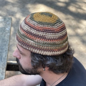 May include: A handmade crocheted beanie hat. The hat features a circular pattern of earthy tones, including olive green, brown, and tan. The beanie has a ribbed brim and a rounded crown. Suitable for all genders.