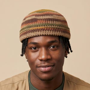 May include: A crochet beanie featuring a horizontal striped pattern in earthy tones of brown, olive green, tan, and rust orange. The hat has a thick, folded brim and a textured, woven construction made from yarn, covering the top of the head.
