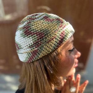 May include: A crocheted beanie hat in a mix of colours including white, green, and burgundy. The hat has a striped pattern and a brim. The hat is shown on a person's head, with the side profile visible. The hat is a handmade item.