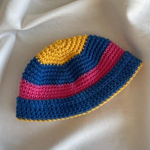 Puede incluir: Un sombrero de pescador tejido a ganchillo con rayas horizontales en tonos vibrantes de amarillo, azul real y rosa intenso. El sombrero está confeccionado con un punto denso y texturizado, con una copa redondeada y un ala corta y elevada.