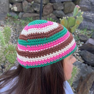 May include: A crochet beanie featuring horizontal stripes in alternating colours of bright green, vibrant pink, cream, and dark brown. The hat has a rounded crown and a fitted silhouette, constructed with a consistent yarn stitch pattern throughout.