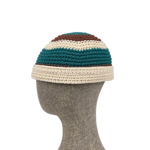 May include: A crocheted beanie hat featuring horizontal stripes in teal, brown, and cream. The hat has a rounded crown and a thick, folded cream-coloured brim. The texture is visible throughout the piece, showcasing the intricate yarn loops.