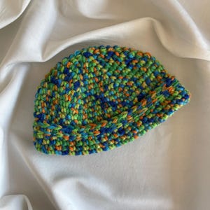 May include: A hand-crocheted beanie hat in a speckled pattern of blue, green, and orange yarn. The brim is folded up. The hat is set against a white background. This handmade hat is a stylish accessory for the colder months.