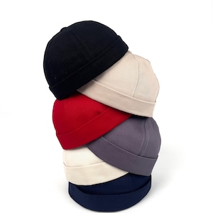 May include: A stack of six solid-colored skull caps in black, beige, red, gray, off-white, and navy blue. The caps are made of a soft fabric and have a short brim. The caps are stacked on top of each other.