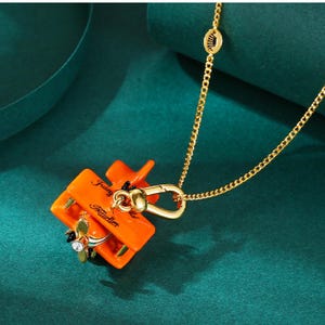 Könnte beinhalten: Orangefarbene Flugzeug-Anhänger-Halskette mit goldfarbener Kette. Der Anhänger trägt die Wörter "Juicy" und "Kouture". Das Flugzeug hat einen Propeller mit einem klaren Kristall und schwarzen Akzenten. Die Halskette befindet sich auf einem tealfarbenen Hintergrund.